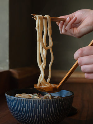 Hands lifting noodles with chopsticks from a bowl of ramen