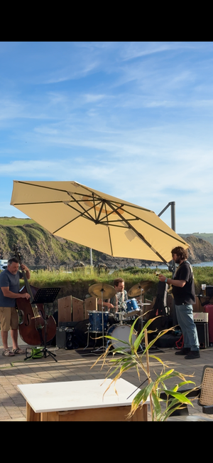 A large beige patio umbrella shading a jazz band performing under a blue sky, with Broad Haven beach in the background